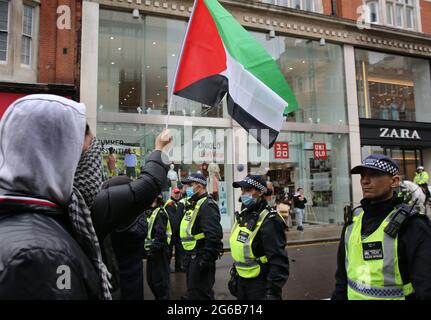 Ein Protestler schwenkt während des Protestes eine palästinensische Fahne auf israelische Unterstützer auf der anderen Seite der Polizeikette.Pro-israelische Demonstranten versammeln sich vor der israelischen Botschaft in der High Street Kensington, um den am 21. Mai vereinbarten Waffenstillstand zu unterstützen. Eine kleine Gruppe von Palästinensern kam zur Gegendemonstration der israelischen Demonstration, doch die Polizei bildete eine Absperrung zwischen den beiden Massen, um jegliche Gewalt zu vermeiden. Stockfoto