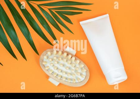 Weiße leere Kosmetiktube mit Creme oder Körperlotion und Holzmassager gegen Cellulite, Palmblatt auf orangefarbenem Hintergrund. Konzept Kampf gegen Cellulite. Stockfoto