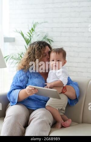 Mutter sitzt zu Hause mit einem Jungen auf dem Sofa, küsst ihn auf der Wange und zeigt einen animierten Cartoon auf einem digitalen Tablet Stockfoto