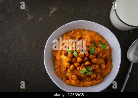 Dicke Kürbissuppe mit gebackenen Kichererbsen und Rettichsprossen auf einem Teller mit einem Glas Milch auf dem Tisch, leckeres gesundes Abendessen Stockfoto