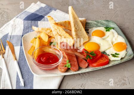 Englisches Frühstück mit Eiern, Würstchen, französischem Toast und Kartoffelkeilen auf einem grauen Tisch Stockfoto