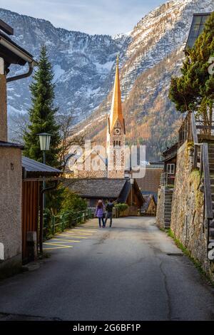 Ein paar Touristen genießen den Blick auf die schöne Hallstatt berühmte Kirche während des Morgens Sonnenaufgang im frühen Frühjahr mit Bergketten in einigen bedeckt Stockfoto