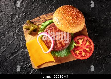 Burger Zutaten, von oben auf schwarzem Hintergrund geschossen. Hamburger Rindfleisch, Cheddar-Käse, Gemüse, Sesambrotbrötchen und Gurken Stockfoto