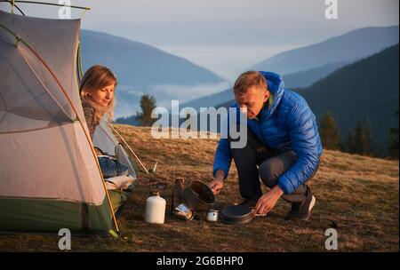 Camping in den Bergen. Wandermann sitzt hockend und gießt Wasser, um Kaffee in der Nähe seiner Frau zu machen, die morgens von ihrem Zelt aus die Schönheit der Umgebung bewundert. Stockfoto