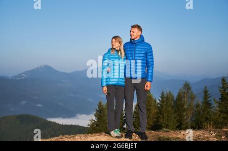 Blick auf die ganze Länge von ein paar Reisenden, während der Spaziergang auf den Bergen hielt oben auf einem von ihnen und Blick nach links, die Landschaft zu genießen. Hinter ihnen wunderschöne Berge und blauer Himmel. Stockfoto
