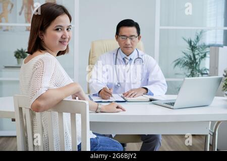 Eine ziemlich lächelnde junge Frau, die am Tisch ihrer Allgemeinpraktizierenden sitzt, die sich Notizen in einem Dokument macht Stockfoto