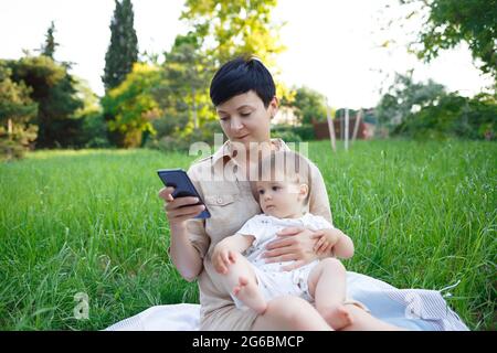 Kleiner Junge, der auf dem Schoß der Mutter sitzt und zum Mobiltelefon zieht. Stockfoto