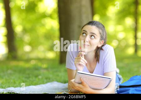 Nachdenkliche Studentin wundert sich, auf die Seite zu schauen, die auf dem Gras liegt und Notizbuch in einem Park hält Stockfoto