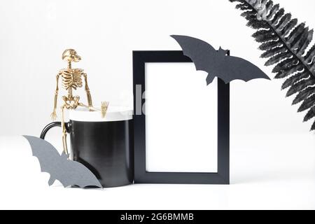 Schwarzer Mockup mit vertikalem Rahmen und schwarzem Becher im Nachbau. Gestylte Stock-Fotografie. Halloween-Konzept mit stilvollen Details Skelett und schwarzen Blättern. Stockfoto