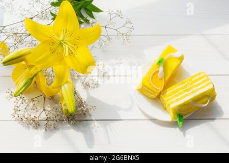 Gelber Kuchen gelbe Lilien auf weißem Holzhintergrund in hellem Sonnenlicht. Blumen Kuchen für den Urlaub. Das Konzept eines Geburtstages, herzlichen Glückwunsch, r Stockfoto