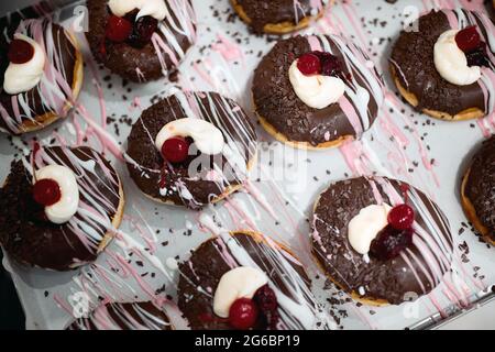 Köstliche Donuts von unwiderstehlichem Aussehen mit Schokoladenüberzug und Kirsche auf dem Tablett in einer Süßwarenwerkstatt. Gebäck, Dessert, süß Stockfoto