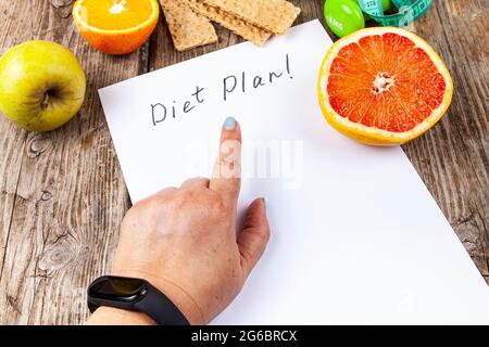 Frau zeigt Finger auf Diät-Plan. Gesunde Ernährung, Kurzhanteln und Maßband auf einem Holzhintergrund. Stockfoto