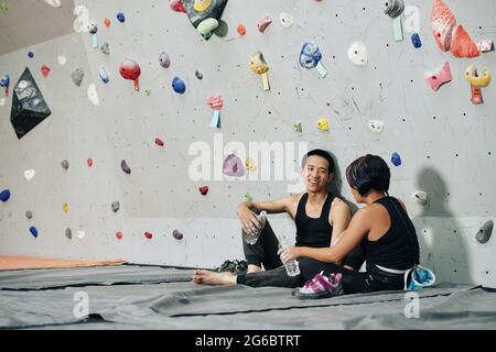 Fröhlicher junger Mann und Frau, die frisches Wasser trinken und reden, wenn sie sich nach dem Klettern im Boulderzentrum ausruhen Stockfoto