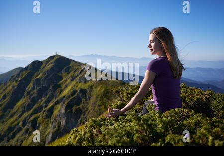 Schöne junge Frau, die auf einem grasbewachsenen Hügel in Lotusposition sitzt und Gyan-Mudra-Handgesten macht, während sie im Freien mediiert. Sportliche, ruhige Frau, die in den Bergen Meditations-Yoga macht. Stockfoto