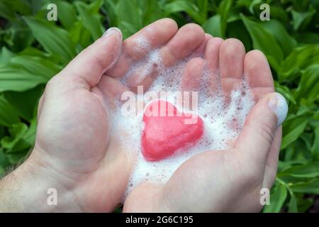 Herrenhände halten rosa Seife mit Schaum in Form eines Herzens auf einem grünen Hintergrund. Die reine Liebe der Männer. Stockfoto