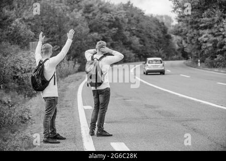 Spüren Sie die Freiheit. Reisen mit Freunden. Fahren Sie mit dem Auto-Stop. Hoffnungsloser Anhalter. Männer versuchen, Auto zu stoppen. Zwillinge gehen entlang der Straße. Wanderer Mann auf der Straße. Weg Stockfoto