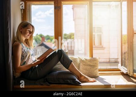 Junge Frau, die zu Hause auf einer gemütlichen, sonnigen Fensterbank sitzt und ein Buch liest Stockfoto