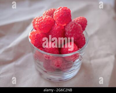 Nahaufnahme von frischen reifen Himbeeren in einem niedrigen transparenten Glas, das auf rauem zerknittertem Papier steht. Stockfoto