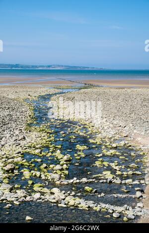 Bach durch Kiesstrand zu Lavan Sands bei Ebbe in der Menai Strait mit Anglesey darüber hinaus ausgesetzt. Llanfairfechan, Conwy, North Wales, Großbritannien Stockfoto