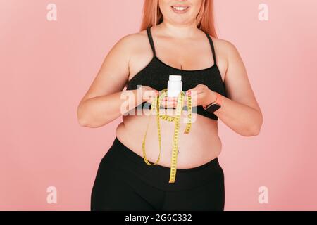 Junge glückliche schöne Frau mit übergroßen Körper trägt Sport-Outfit hält Pille Pack und Maßband, während auf rosa Hintergrund. Stockfoto