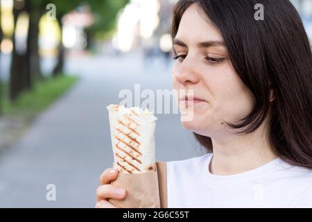 Brünette Frau überfrisst Shawarma auf einer Straße in der Stadt. Street Fast Food Pita Roll mit Fleisch und Gemüse. Stockfoto
