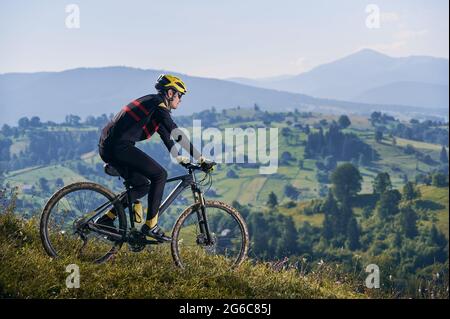 Horizontale Momentaufnahme eines Mannes, der am frühen nebligen Morgen mit seinem Fahrrad in den Bergen fährt und bergab fährt. Gipfel in Wolken, grüne Hügel im Hintergrund. Seitenansicht, Kopierbereich. Konzept des aktiven Lebensstils Stockfoto