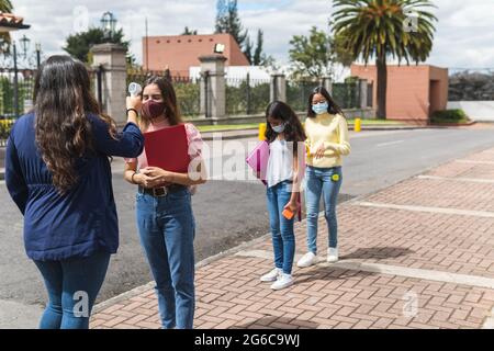 Eine Lehrerin mit dem Rücken drehte sich um, um die Temperatur mit einem Infrarot-Thermometer einer jungen Latina-Schülerin zu messen, während zwei Klassenkameraden auf sie warteten Stockfoto