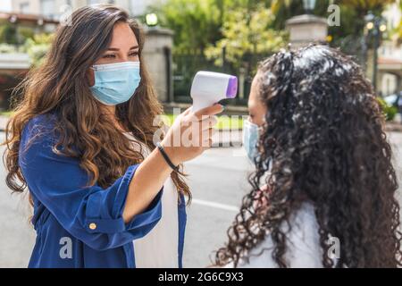 Selektiver Fokus eines Latina-Lehrers, der eine Maske trägt und die Temperatur eines Teenagers mit einem Infrarot-Digitalthermometer überprüft. Zurück zur Schule Stockfoto