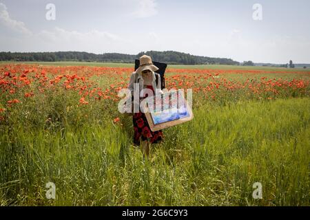 Reife Künstlerin malt ein Bild auf einem Feld blühender Mohnblumen, die die Künstlerin aus der freien Luft zurückkehrt und das gemalte Bild trägt Stockfoto