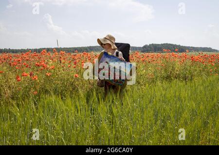 Der Künstler kehrt aus dem Freien zurück und trägt das gemalte Bild auf einem Feld blühender Mohnblumen Stockfoto