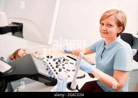 Sonografin mit sterilen Handschuhen, die weibliche Patientin mit einem Ultraschallgerät untersucht. Ärztin, die die Kamera anschaut und lächelt, während sie eine Ultraschalluntersuchung macht. Konzept der Ultraschalldiagnostik. Stockfoto