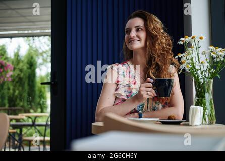 Charmante Frau mit lockigem Haar, die Kaffee trinkt, sitzt am Tisch im gemütlichen Kaffeehaus Stockfoto