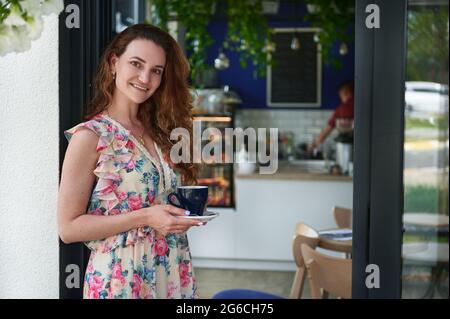 Junge lächelnde Frau, die mit einer Tasse Kaffee in den Händen am Eingang ihrer Cafeteria steht. Geschäftsfrau, Unternehmerin, Cafeteria-Inhaberin Stockfoto