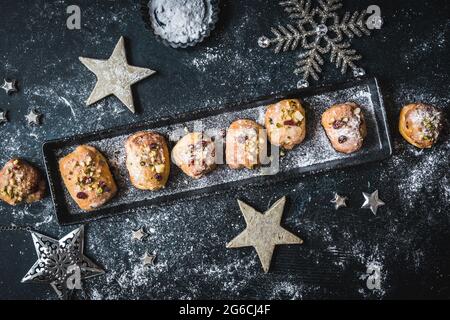 Hausgemachter Mini-Stollen mit Sahnehäubchen, getrockneten Preiselbeeren und Pistazien auf einem schwarzen Tablett auf schwarzem Hintergrund, verziert mit silbernen Sternen. Draufsicht. Stockfoto