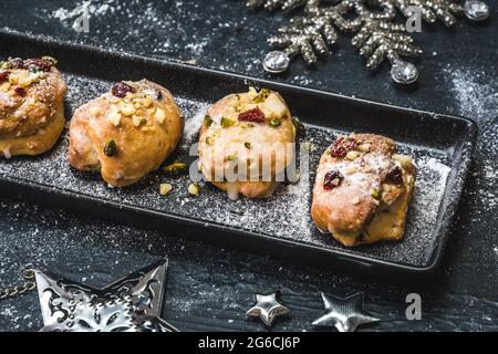 Hausgemachter Mini-Stollen mit Sahnehäubchen, getrockneten Preiselbeeren und Pistazien auf einem schwarzen Tablett auf schwarzem Hintergrund, verziert mit silbernen Sternen. Stockfoto