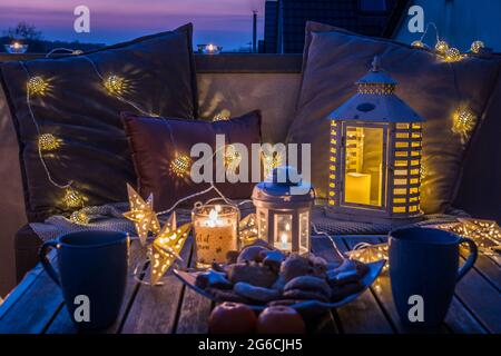 Gemütliche Sitzecke mit Kaffee und Keksen auf dem Balkon, dekoriert für den Winter mit Laternen, Kerzen, Wolldecken und Lichterketten Stockfoto