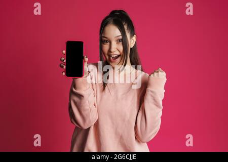 Asiatische aufgeregt Frau zeigt Gewinner Geste und Handy isoliert über lila Wand Stockfoto