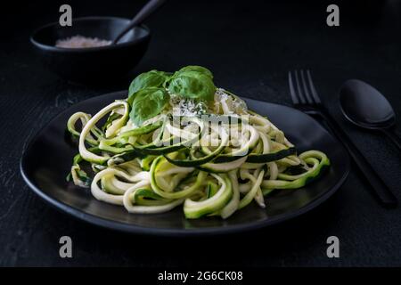 Zoodles, Zucchini-Nudeln mit Parmesan und Basilikum auf einem schwarzen Teller auf schwarzem Hintergrund. Für Low Carb, Keto, Vegetarier, Paläo Ernährung und Ernährung. Stockfoto