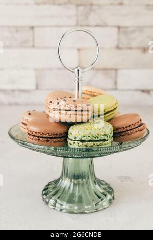 Bunte französische Macarons auf einer Glastagere vor einer weißen Wand. Gebäck, Desserts und Süßigkeiten. Vertikales Stockfoto. Stockfoto