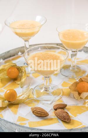 Drei Gläser mit Eierwein, Physalis-Früchten und gesalzenen Mandeln mit Schale auf einem runden Tablett auf weißem Grund, vertikal Stockfoto