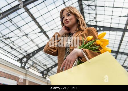 Blick aus der unteren Perspektive auf eine junge Frau mit einer Sonnenbrille, die gelbe Tulpen und Einkaufstaschen hält Stockfoto