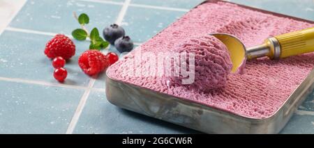 Leckeres rosafarbenes Beereneis oder Sorbet im Topf mit Löffel, der mit frischer Himbeere und roter Johannisbeere und Heidelbeere auf den Tisch gelegt wird Stockfoto