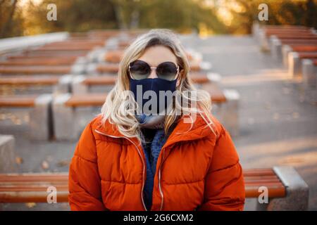 Blonde Frau in Oberbekleidung und Maske zu Fuß auf gepflasterten Weg an sonnigen Tag im Herbst Park während Pandemie Stockfoto
