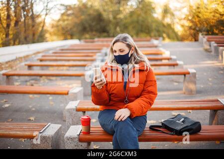 Blonde Frau in Oberbekleidung und Maske zu Fuß auf gepflasterten Weg an sonnigen Tag im Herbst Park während Pandemie Stockfoto