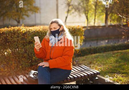 Blonde Frau in Oberbekleidung und Maske zu Fuß auf gepflasterten Weg an sonnigen Tag im Herbst Park während Pandemie Stockfoto