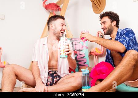 Lustig aufgeregt verschiedene Erwachsene homosexuelle Freunde in Sommer-Outfits mit Getränken, die vorgeben, am Strand mit rosa Flamingo und Spaß zusammen Stockfoto