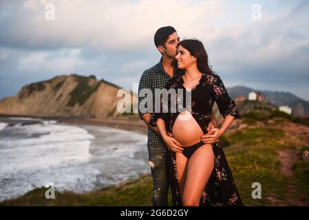 Liebevoller Mann umarmt und küsst glücklich schwanger Frau in stilvollem Outfit enthüllt Bauch gekleidet, während zusammen am Meer gegen Berge stehen Stockfoto