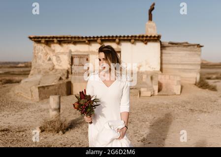 Frau in weißem Kleid und mit Blumen, die am Hochzeitstag im Naturpark Bardenas Reales in Navarra, Spanien, gegen eine heruntergekommene Hütte stehen Stockfoto