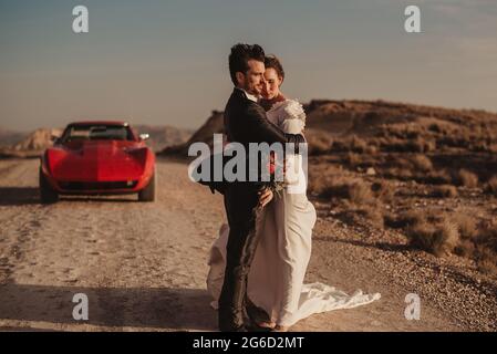 Seitenansicht der Braut und des Bräutigams, die sich umarmen, während sie bei einem roten Luxusfahrzeug während einer Fahrt durch den Naturpark Bardenas Reales in Nava stehen Stockfoto