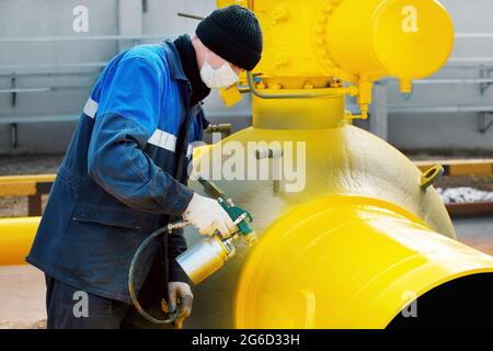 Ein Maler in Arbeitskleidung malt an einem Sommertag ein Absperrventil aus Metall zur Vergasung aus einer Kompressorpistole. Professionelles Lackieren von Teilen. Industrieller Hintergrund. Stockfoto